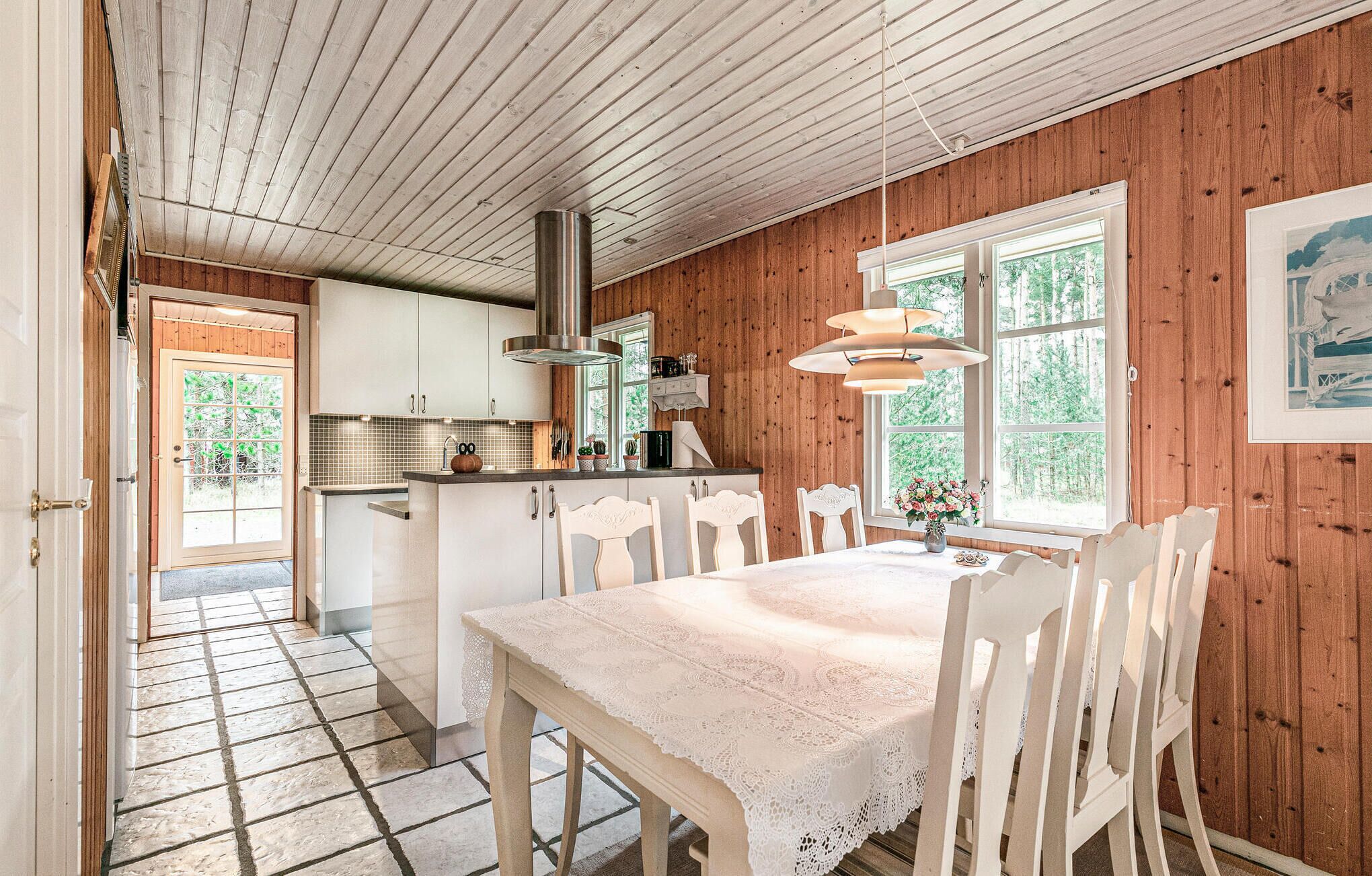 A cozy kitchen with wooden walls, a dining area with a lace tablecloth, and a large window with a view of trees.