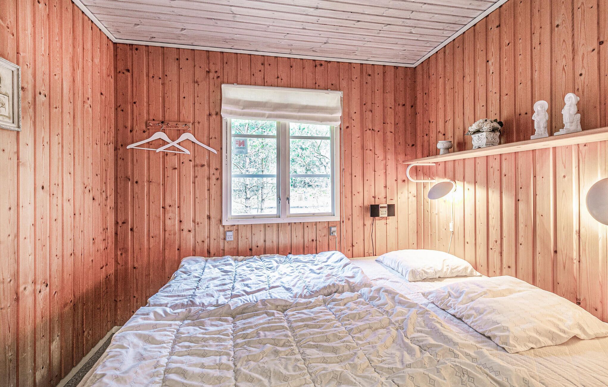 A bedroom with wooden walls, a bed with white bedding, a window with a view of trees, and a shelf with decorative items.