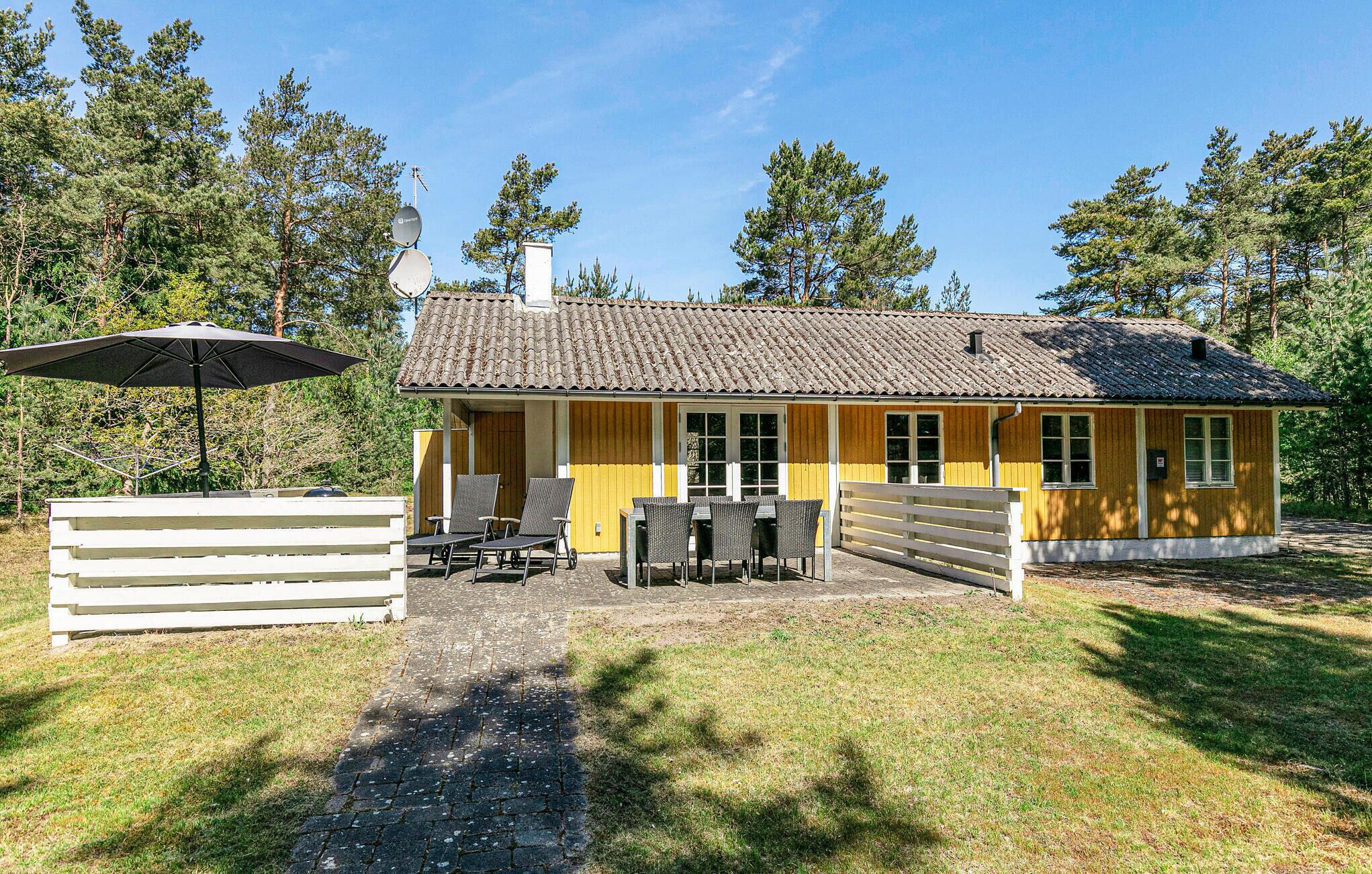 A yellow building with a tiled roof, a covered patio with chairs and an umbrella, and a white fence in front.