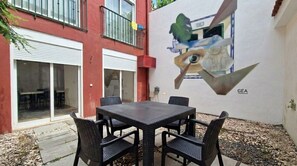An outdoor patio with a table and chairs, a mural on the wall, and a building with a red facade and a balcony.
