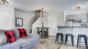 A modern living room with a grey sofa, red and black plaid throw pillows, a wooden coffee table, and a staircase leading to a kitchen area with bar stools.