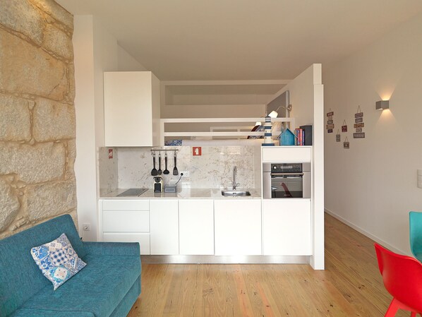 A compact kitchen with white cabinets, a marble countertop, and a built-in oven. There's a teal sofa with a patterned pillow and a red chair in the living area.