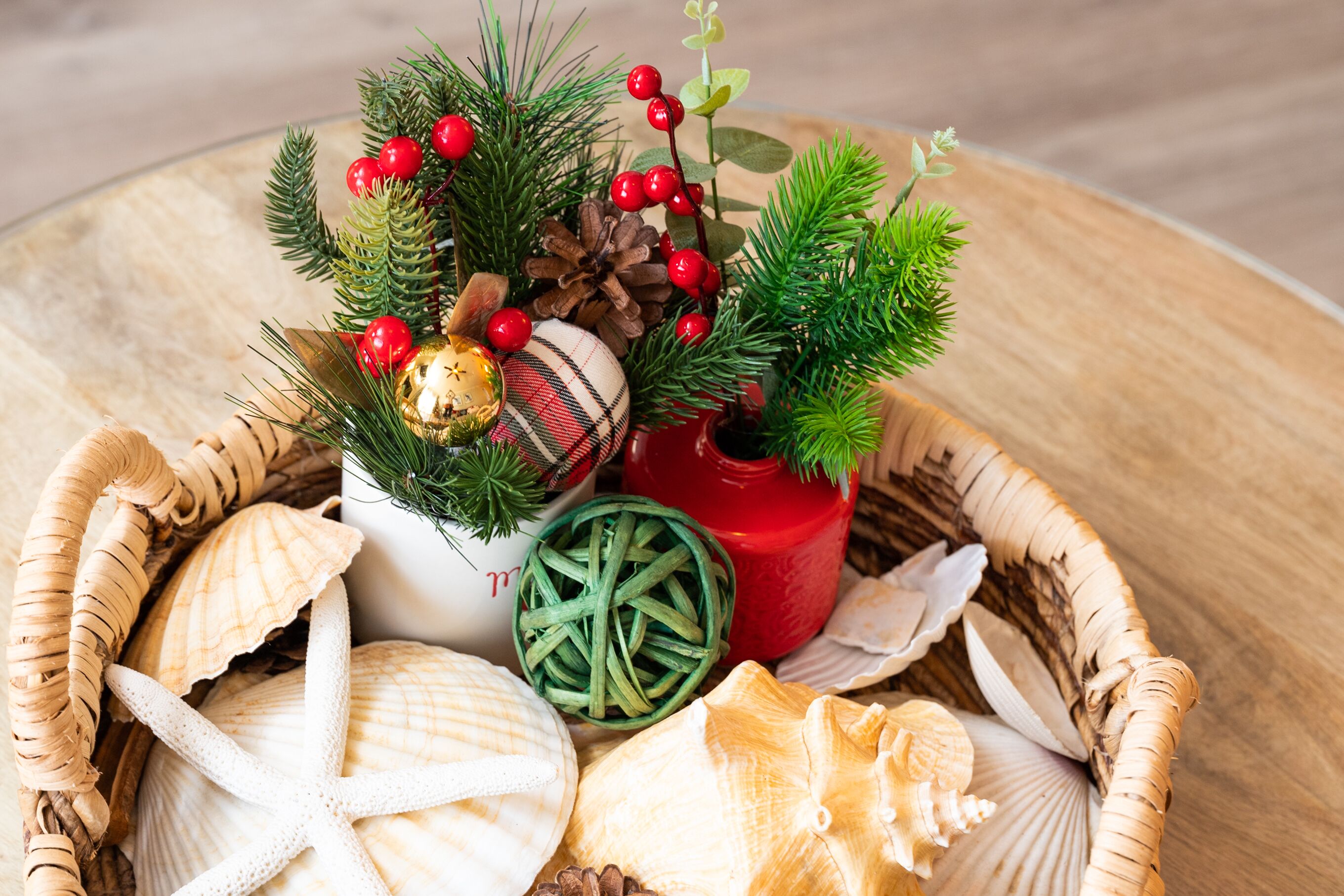 A wicker basket containing seashells, pine branches, red berries, and a decorative pine cone.