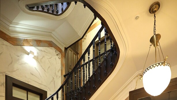 Una imponente escalera en espiral con peldaños de madera y barandillas negras, que conduce a un descansillo con un espejo grande y decorativo y una araña.