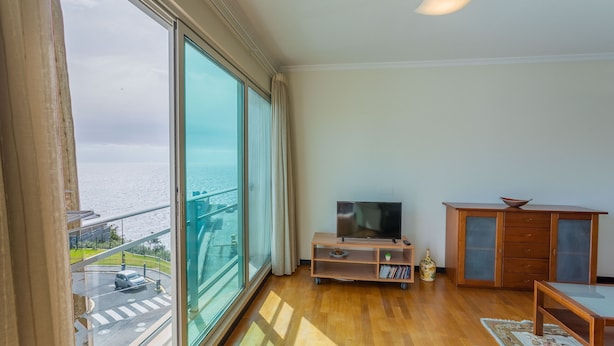 Habitación con un amplio ventanal con vista a un cuerpo de agua, un televisor sobre un soporte de madera, un mueble con cajones y una pequeña mesa.