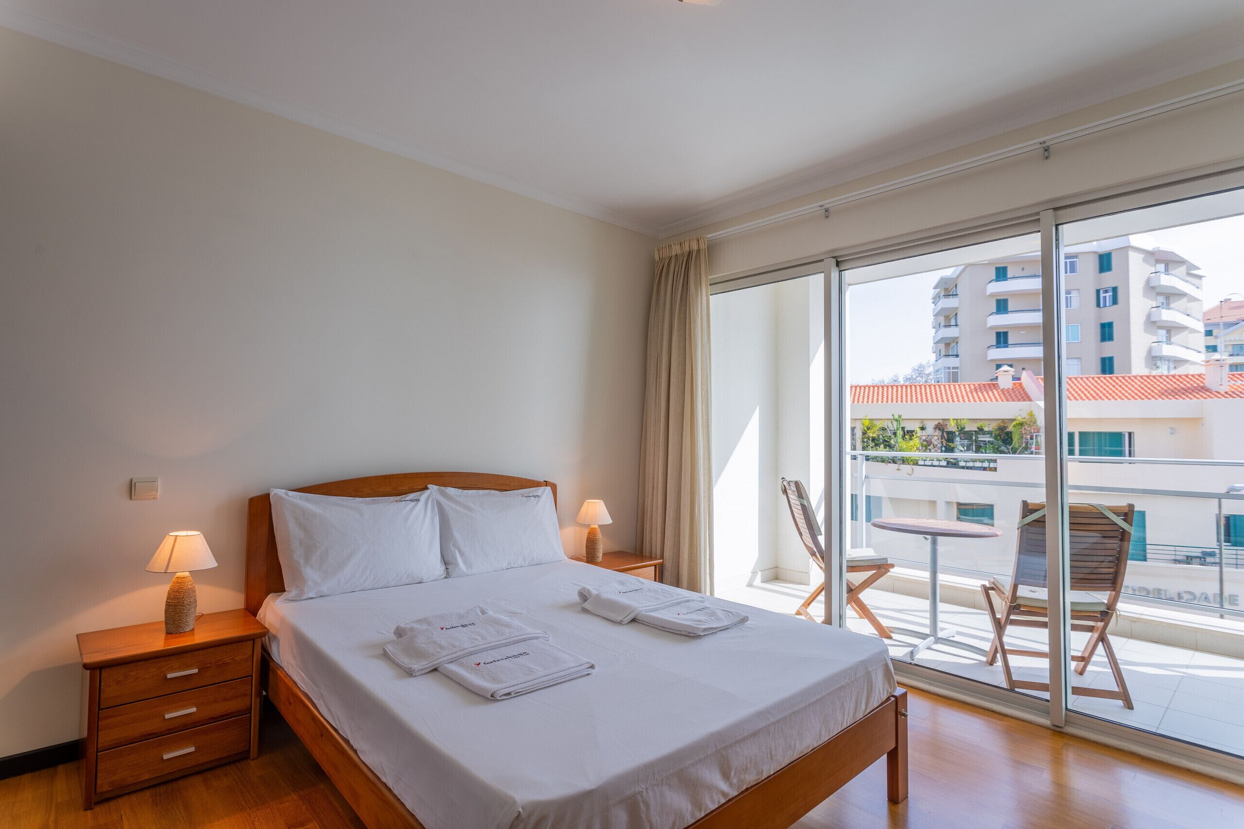 A hotel room with a bed, a nightstand with a lamp, a window with a view of buildings, and a balcony with a table and chairs.