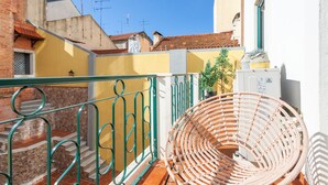 Un balcon avec un fauteuil en osier, une rambarde en métal et une vue sur les immeubles voisins.