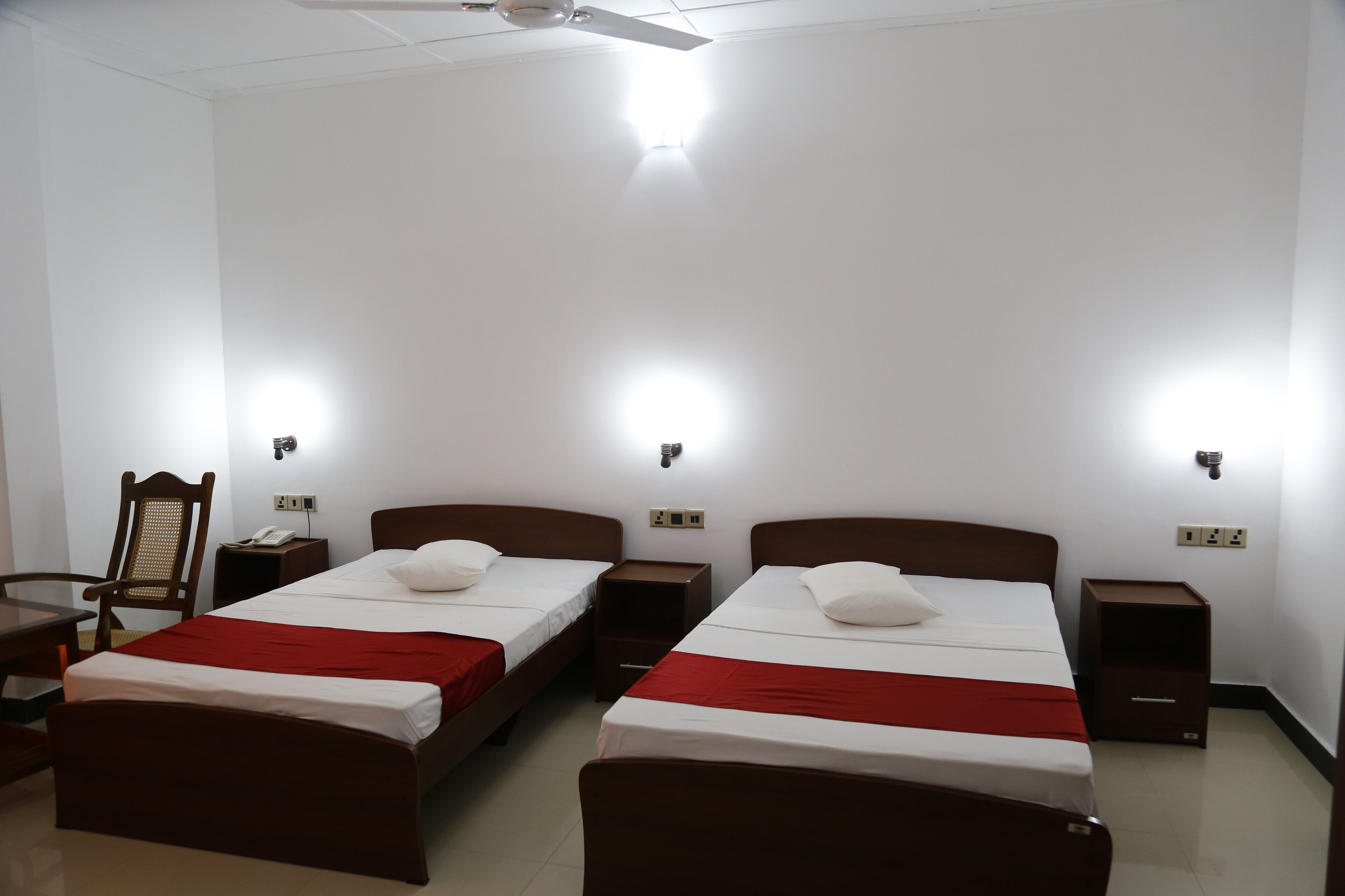 Habitación de hotel con dos camas, cada una con una colcha roja y almohadas blancas. Hay mesitas de noche, una silla y un ventilador de techo.