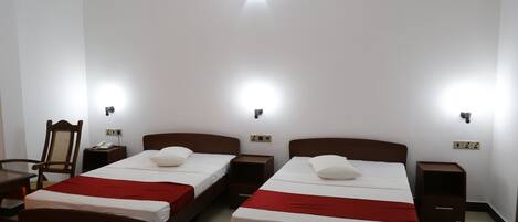 Habitación de hotel con dos camas, cada una con una colcha roja y almohadas blancas. Hay mesitas de noche, una silla y un ventilador de techo.