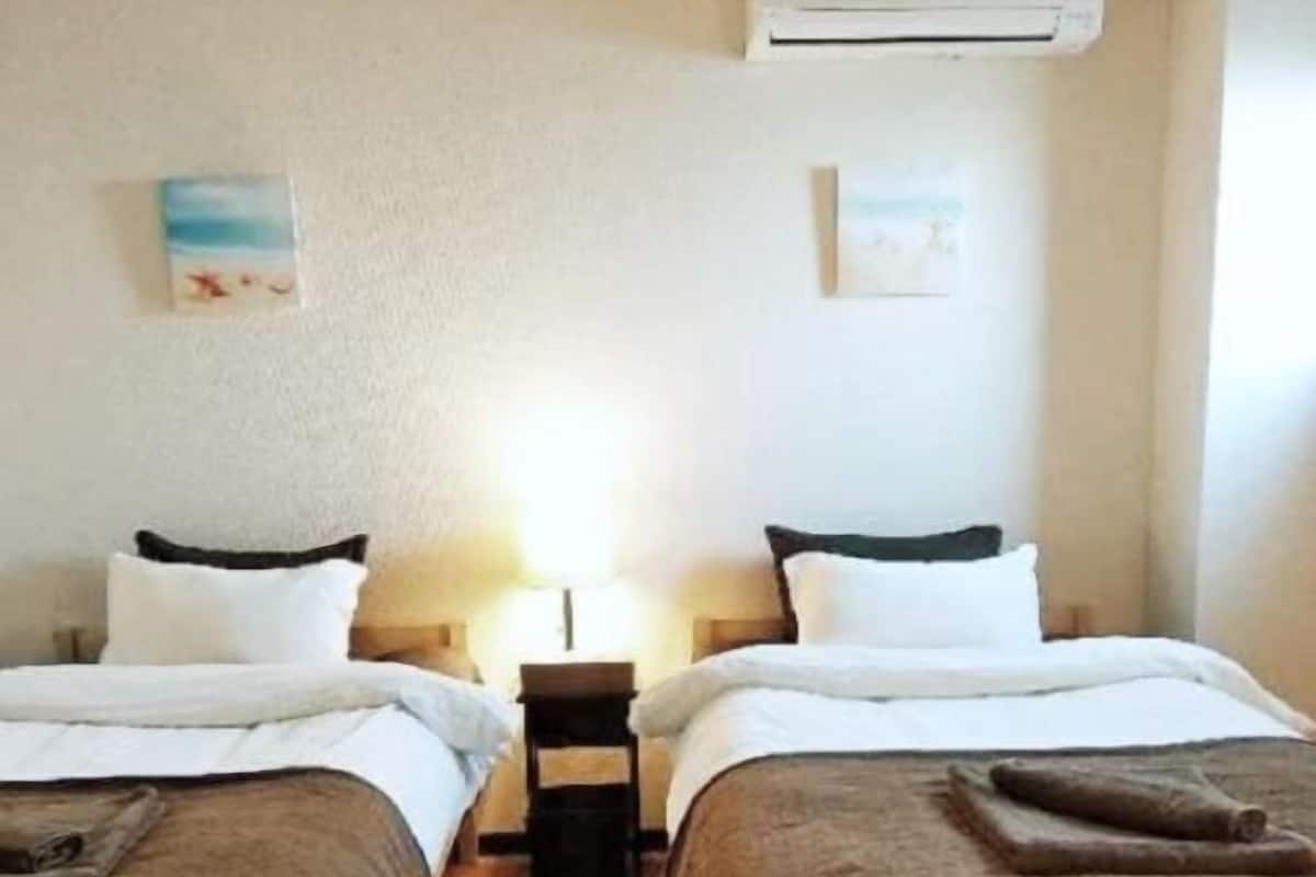 A hotel room with two single beds, a nightstand, an air conditioning unit, and a small table with a lamp.