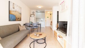 Una sala moderna con sofá, sillas, una mesa de centro y televisión. Se observa una cocina al fondo con armarios y un microondas.