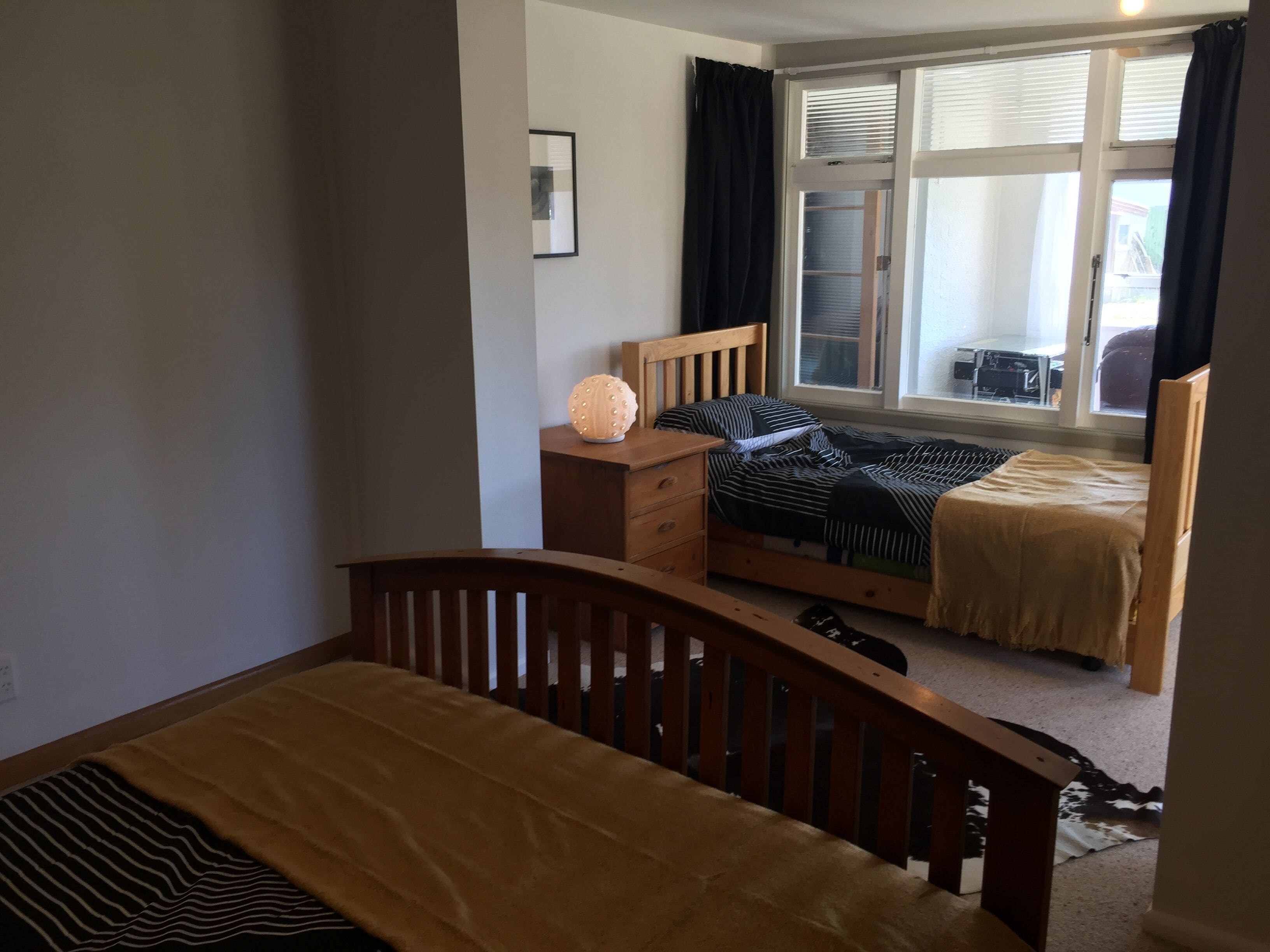 A bedroom with a single bed, a wooden dresser, a bedside table with a lamp, and a window with curtains.