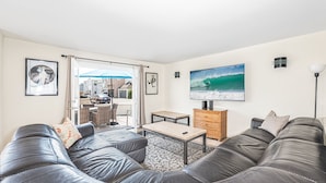 Una sala de estar con un sofá grande de color gris, una mesa de centro y un televisor en la pared. Se observa un cuadro con una imagen de un surfista y una escena de playa.