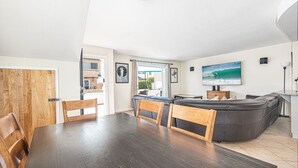 Una sala moderna con piso de madera oscura, un sofá de cuero negro, sillas de madera, una mesa de comedor y un televisor que muestra una escena de surf.