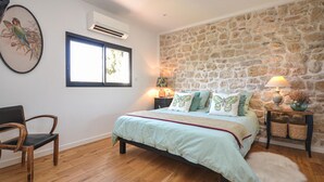 Un dormitorio con pared de piedra, una cama con varios almohadones, una silla, una mesita de noche con lámpara y un equipo de aire acondicionado.