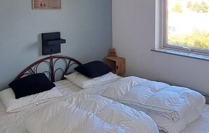 Une chambre à coucher avec un lit, des oreillers, une table de chevet, un ours en peluche et une fenêtre.
