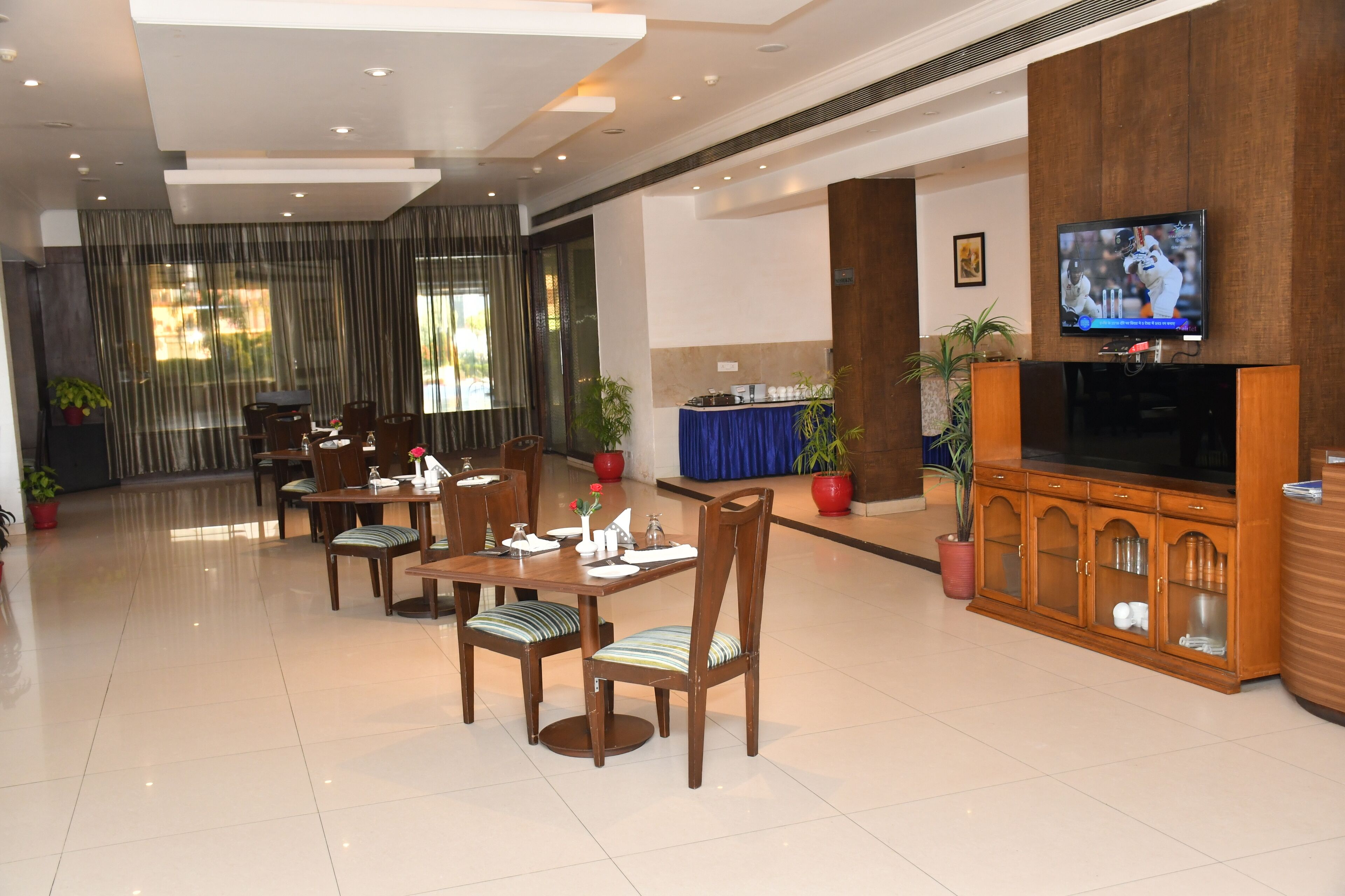 A hotel lobby with a dining area, a television mounted on a wooden cabinet, and potted plants.