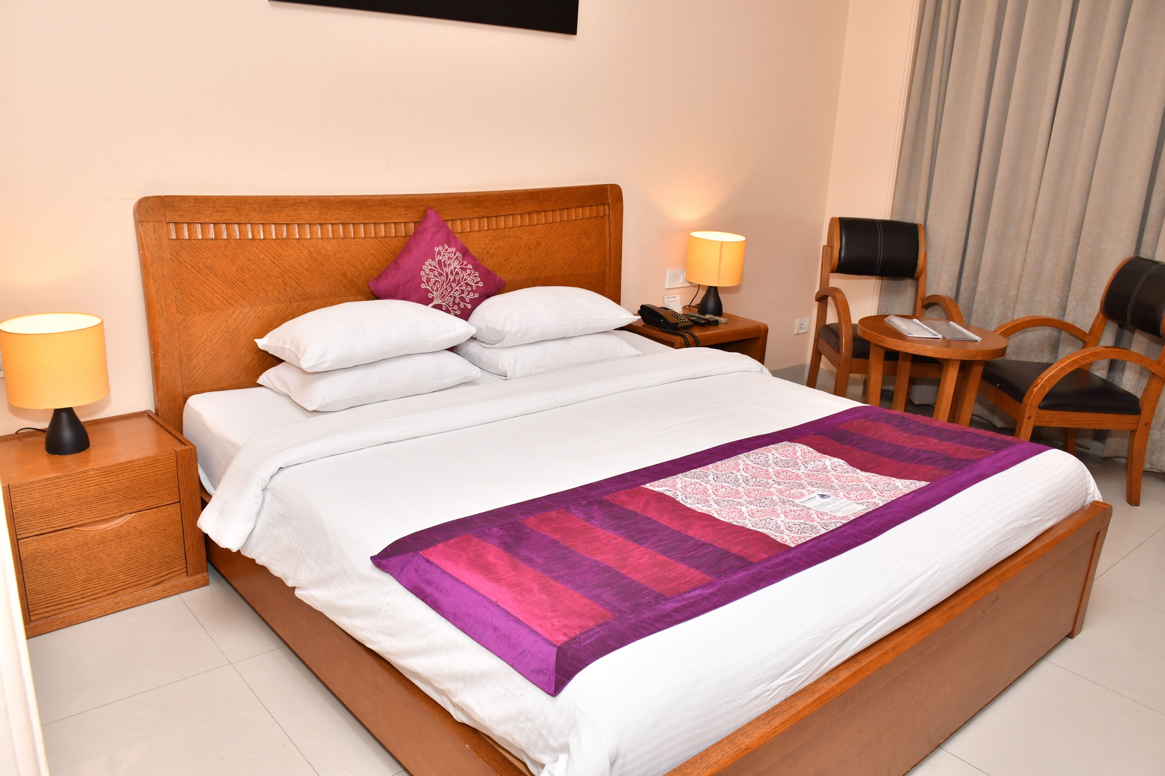A neatly made bed with white linens and a purple and pink quilt, a wooden nightstand with a lamp, a small table with a lamp, and a chair.