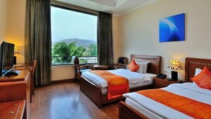 Habitación de hotel con dos camas, un escritorio, una silla y un ventanal con vistas a zonas verdes y colinas.