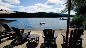 Uitzicht op een meer met ligstoelen en een tafel onder een parasol, een zeilboot op het water, en een beboste heuvel op de achtergrond.
