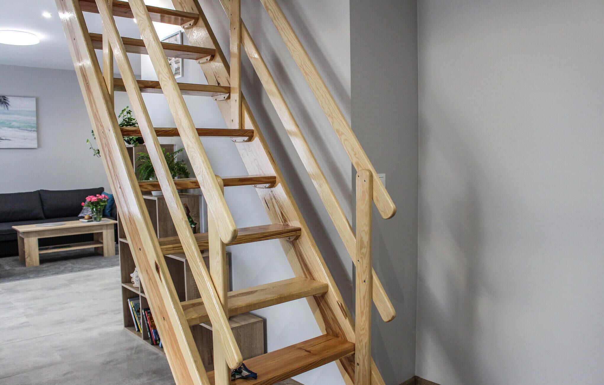 A wooden ladder leaning against a wall in a room with a gray floor, a gray wall, and a gray couch in the background.