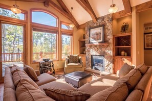 A cozy living room with a stone fireplace, large windows, and a wooden ceiling.