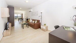 A modern living room with a brown sofa, a coffee table, and a TV on a wooden stand. There is a dining area in the background with a table and chairs. - Summer Breeze Comfort Apartment (Il-Mellieha)