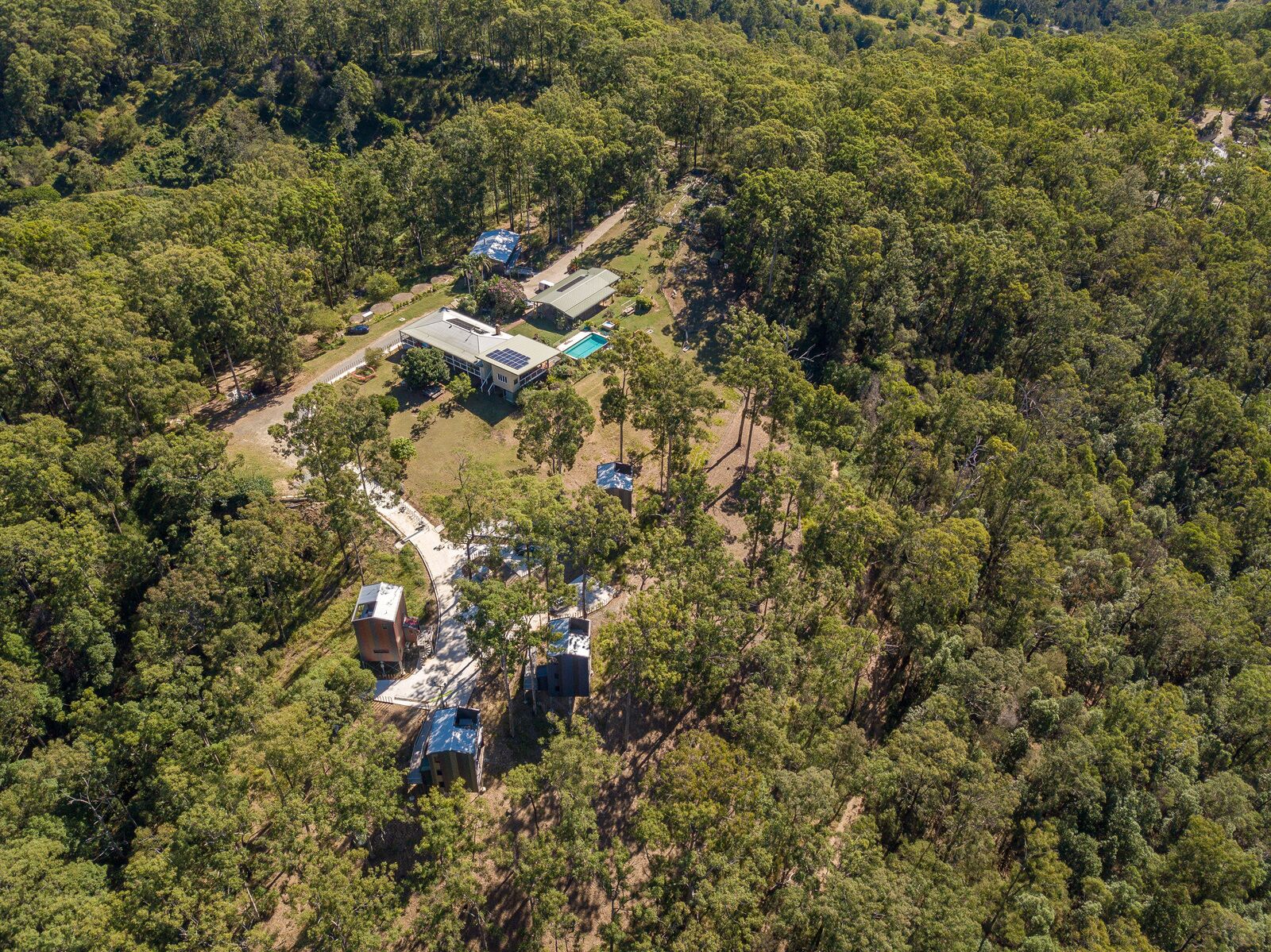 Vue aérienne d’une propriété comprenant plusieurs bâtiments, entourée d’une forêt dense. On y distingue différentes constructions, notamment un grand bâtiment doté d’un toit vert et des bâtiments plus petits avec des toits bleus.