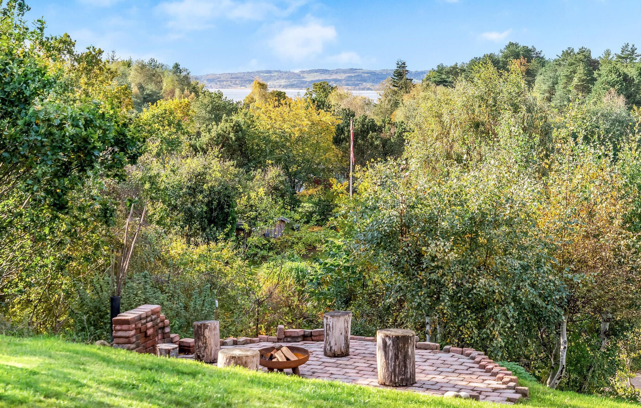 Un espacio de estar circular al aire libre con un brasero, rodeado por un muro de ladrillo y postes de madera, situado en un exuberante jardín con árboles y una vista distante de colinas.