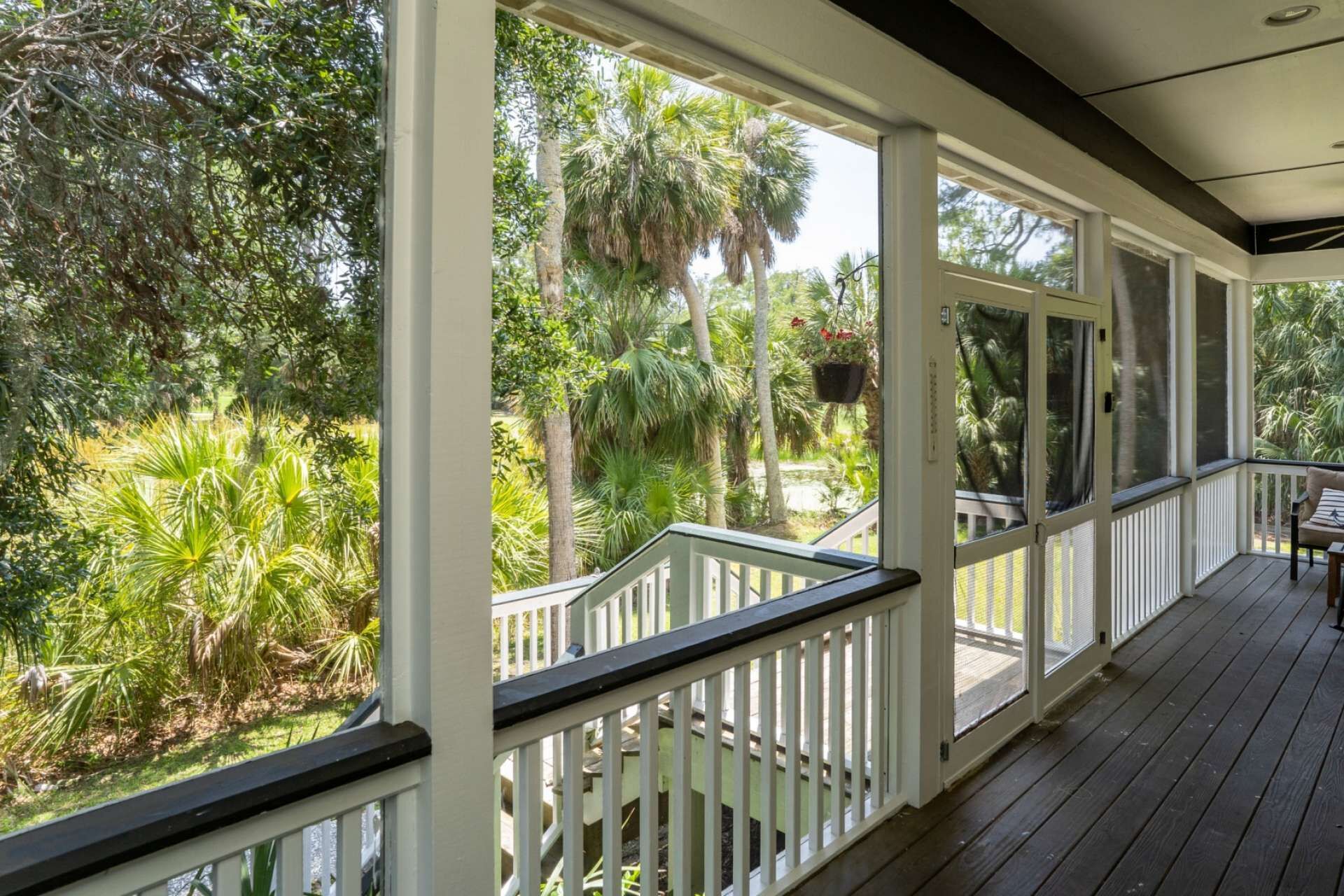 A covered porch with white railings and a wooden deck, overlooking a lush garden with various tropical plants and trees.