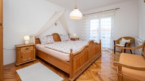 Quarto com cama de madeira, mesa de cabeceira, cadeira e janela com cortinas leves.