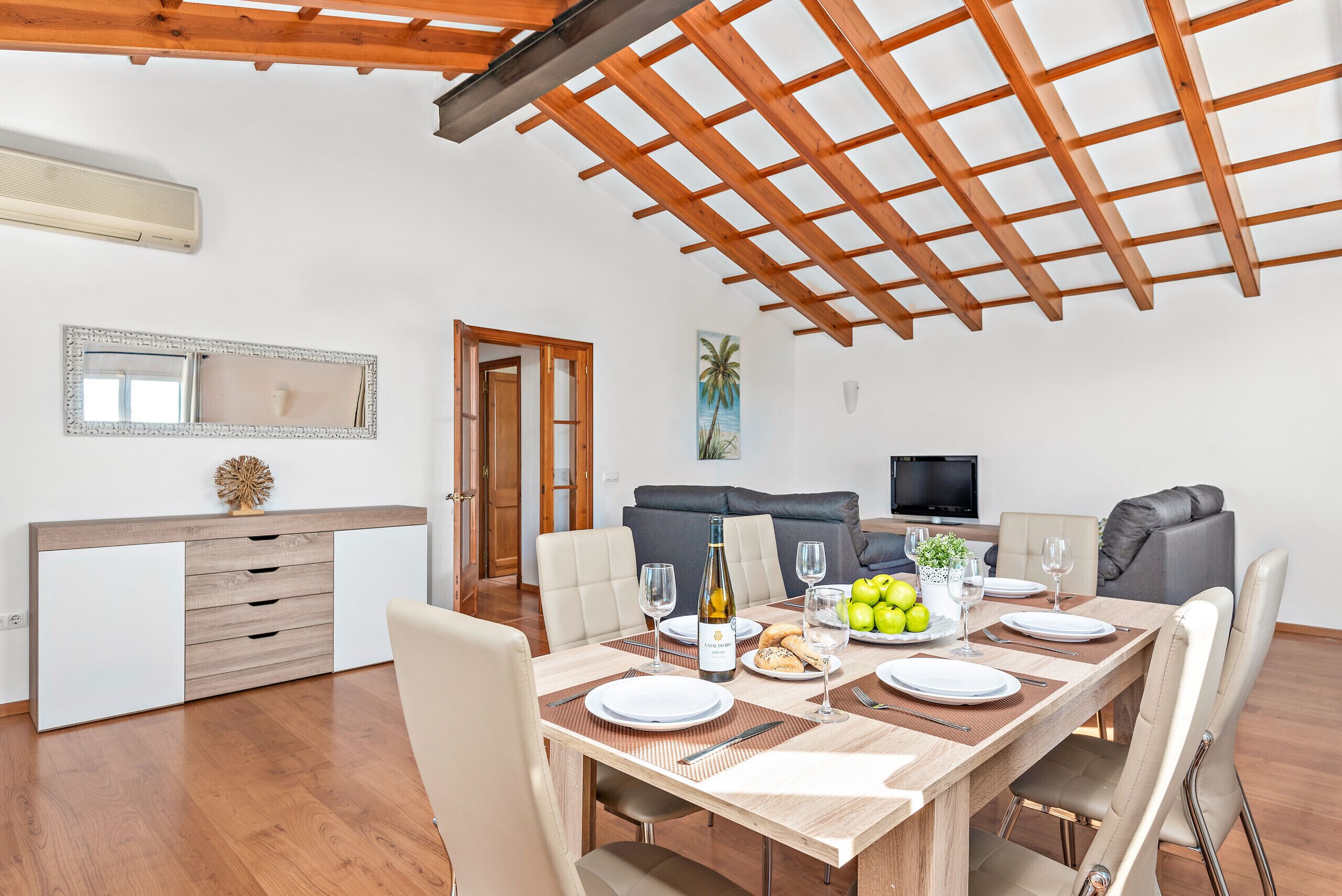 Un comedor con una mesa de madera dispuesta para cuatro personas, un sofá, un televisor y un cuadro en la pared.