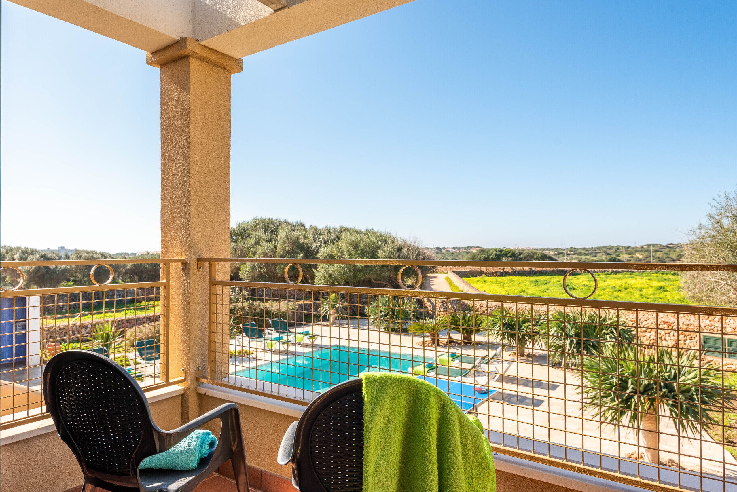A balcony with two chairs, one with a green towel, overlooking a swimming pool and a landscaped garden.