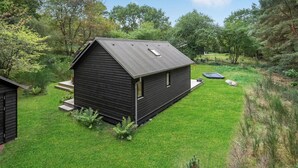 A small, dark wooden cabin with a sloped roof, surrounded by lush green grass and trees. - Stunning home in Fårevejle with kitchen (Fårevejle)