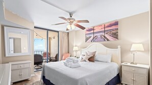 A hotel room with a large bed, a ceiling fan, a painting of a beach, a desk, and a chair.