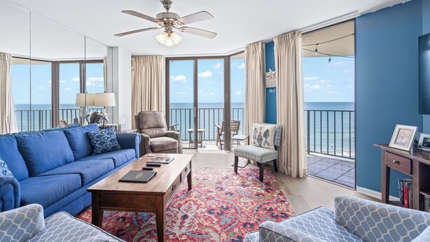 A spacious living room with a blue sofa, a wooden coffee table, and a patterned rug. There is a ceiling fan, a large window with a view of the sea, and a balcony with a table and chairs.