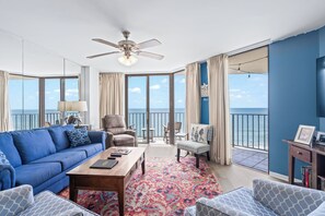 A spacious living room with a blue sofa, a wooden coffee table, and a patterned rug. There is a ceiling fan, a large window with a view of the sea, and a balcony with a table and chairs.