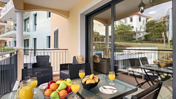 A balcony with outdoor furniture, a table with fruit and drinks, and a view of buildings and greenery. - Apartment Lotus by LovelyStay (Funchal)