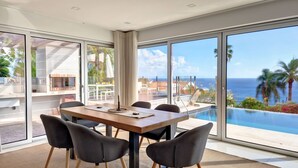 Un espace de restauration moderne doté d’une grande table et de chaises, ainsi que de larges fenêtres offrant une vue sur l’océan et les palmiers.