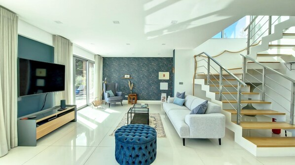 Una sala de estar moderna con una televisión de pantalla plana, un sofá gris, un reposapiés azul y una escalera con escalones de madera.