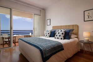 A hotel room with a bed, a chair, a table, and a view of the ocean through a sliding door. - Vista Monumental - Three Bedroom Apartment, Sleeps 6 (Funchal)