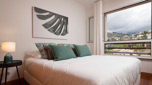 Un dormitorio con una cama grande, una mesita de noche con lámpara y una ventana que da a vista de un pueblo.