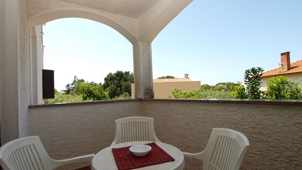 A covered outdoor seating area with a round table and four white chairs, a red placemat, and a view of greenery and buildings in the background.