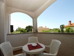 Un espacio de estar al aire libre cubierto con una mesa redonda y cuatro sillas blancas, un mantel individual rojo, y vistas a zonas verdes y edificios al fondo.