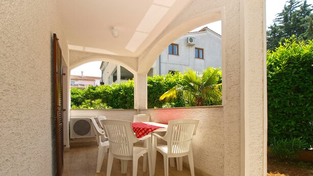 A covered outdoor seating area with a table and chairs, air conditioning units, and a view of greenery and buildings in the background.