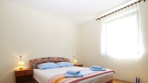 A bedroom with a wooden bed, two bedside tables with lamps, a window with sheer curtains, and a wooden floor.