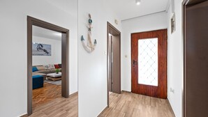 Un corridoio con pavimento in legno, una porta in legno con finestra a motivi a diamante, e un soggiorno con divano e tavolino.