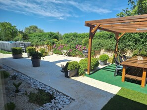 A garden patio with a wooden pergola, potted plants, a barbecue grill, and outdoor seating.