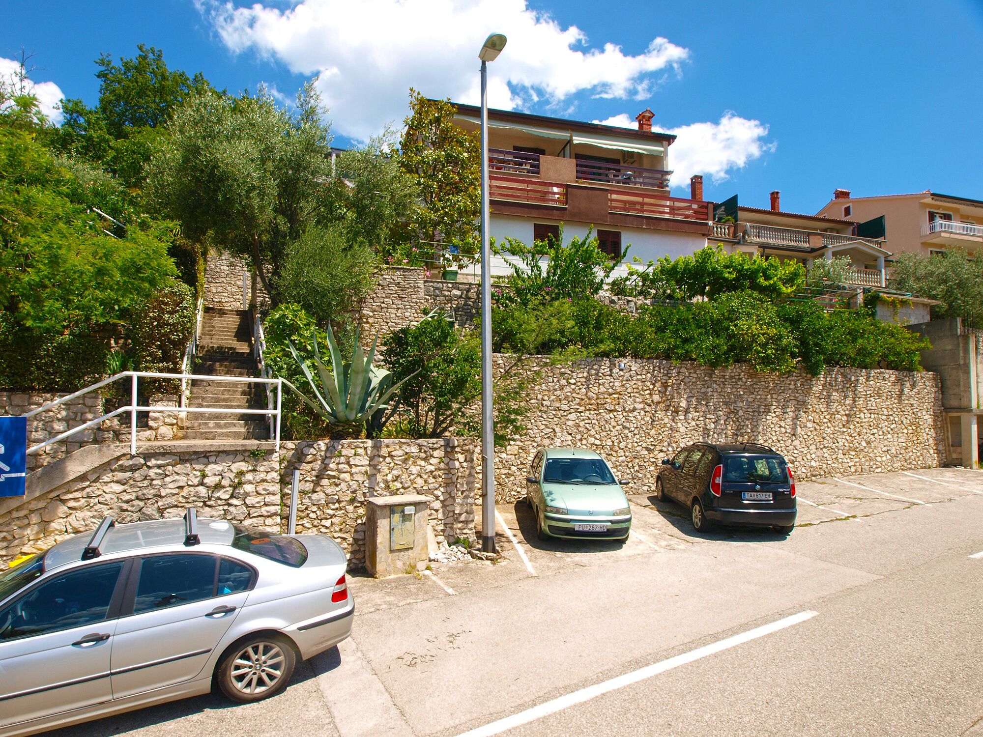 Una zona de pàrquing amb dos cotxes, una paret de pedra i una escala que porta a un edifici amb balcons.