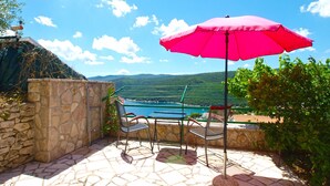 Une terrasse avec un sol en pierre, deux chaises en métal, un parasol rouge et une vue sur un lac et des collines.